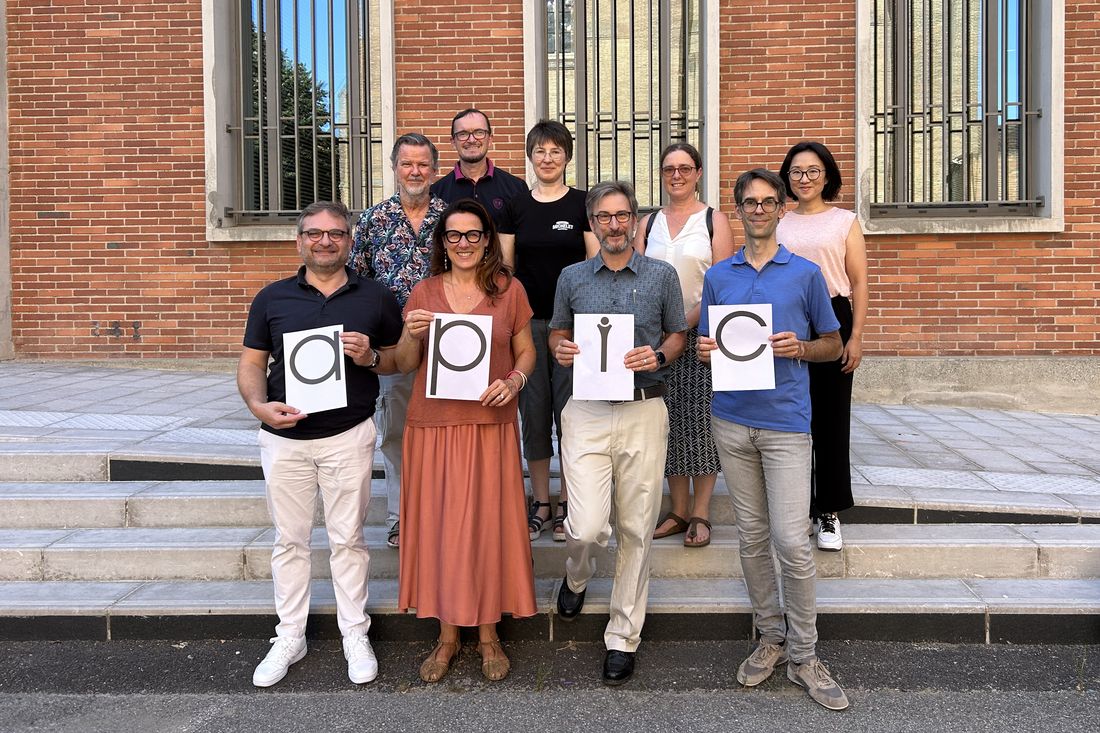 Les représentants de l'APIC réunis devant une façade de brique toulousaine ; quatre d'entre eux tiennent des feuilles formant le mot « apic »